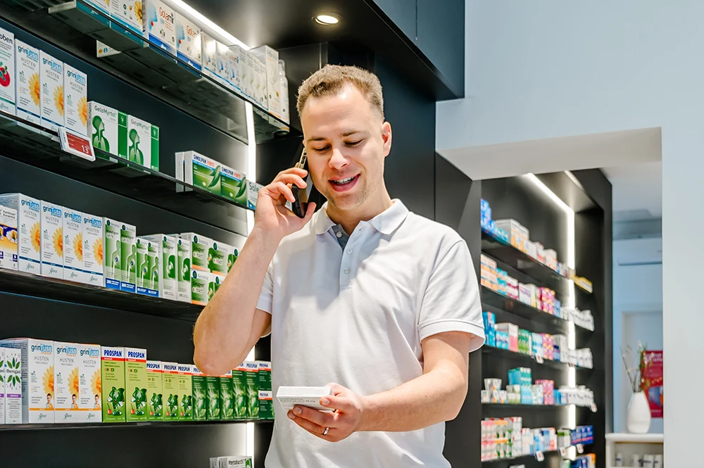 Ein Mann im weißen Poloshirt steht lächelnd in einer Apotheke und berät eine Kundin telefonisch zu einem Rezept und Botendienst. Er hält eine Medikamentenpackung in der Hand und blickt auf Regale voller Medikamentenpackungen.