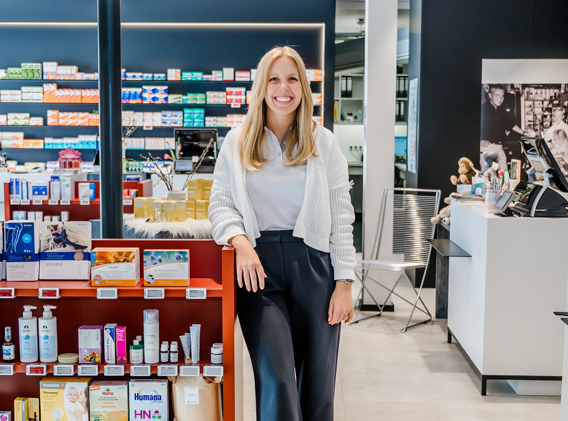 Eine lächelnde Apothekerin steht in einer modernen Apotheke und lehnt an einem Regal mit Gesundheitsprodukten und Medikamenten. Bunte Verpackungen säumen die Regale, im Hintergrund ist der Apothekentresen zu sehen.